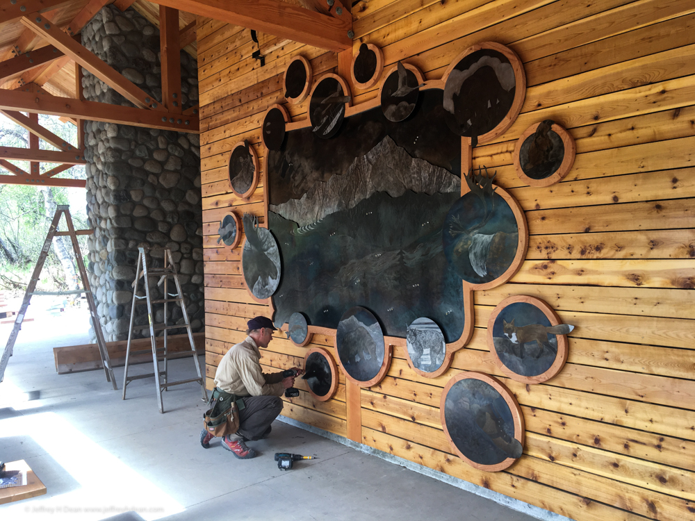 Final installation steps for 'Through Your Spotting Scope' at Kesugi Ken Interpretive Center in Denali National Park. Large outdoor metal wall mural with curved wood frame